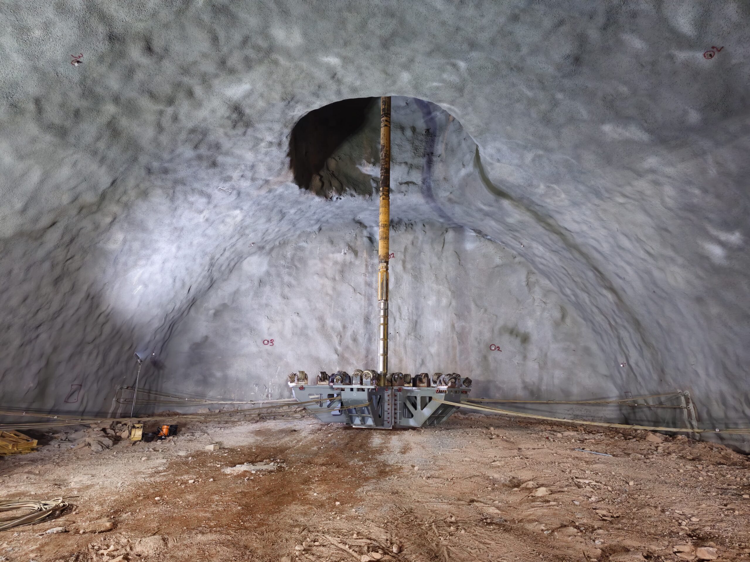 Large underground space with curved, textured walls. A cylindrical metal structure extends from floor to ceiling, secured with cables. The area is dimly lit, and the floor is covered with dirt and rocks. Construction equipment is visible.