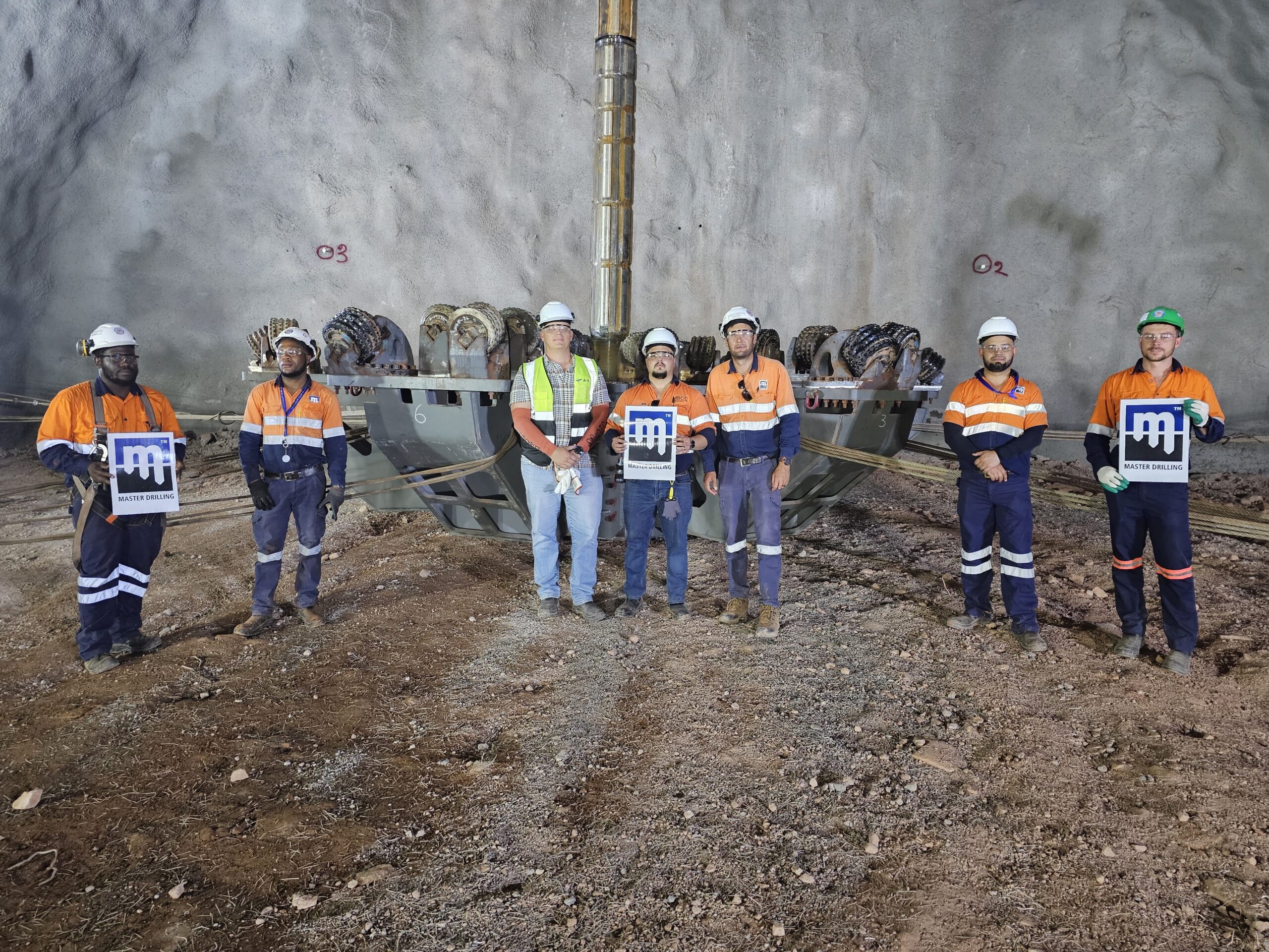 Six construction workers in safety gear stand in front of a large tunnel boring machine. They hold company logos and wear high-visibility clothing, helmets, and gloves in a rocky underground environment.