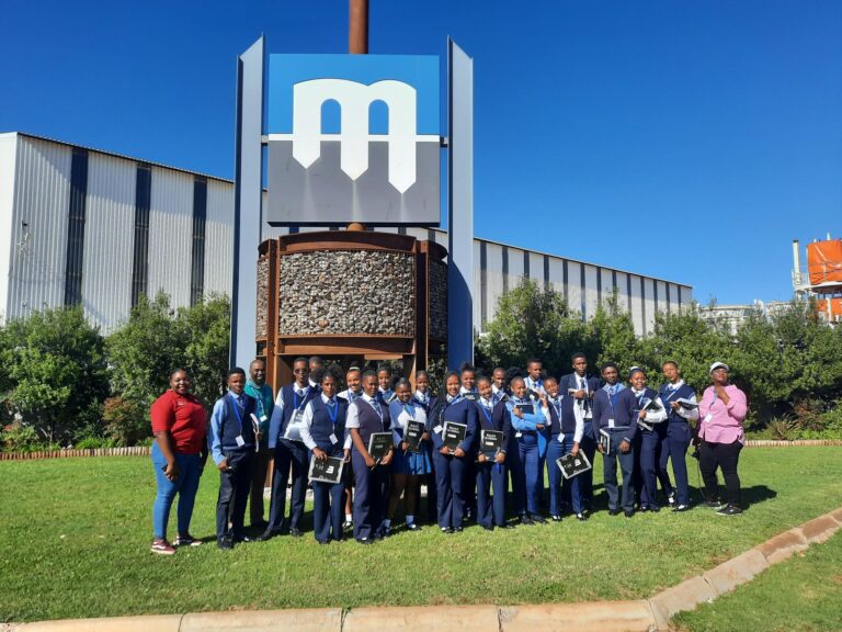 A group of students in blue and white uniforms stands in front of a large industrial building with a sign featuring a large "M." Two adults stand at the edges of the group, posing for a photo under a clear blue sky.