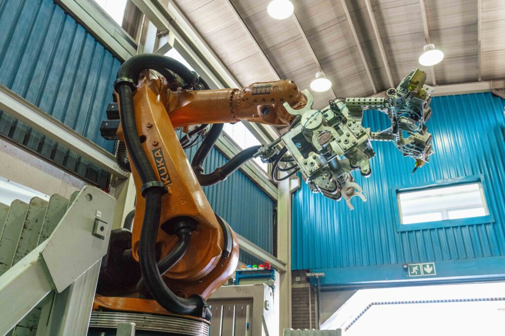 A large industrial robotic arm with a complex gripper attachment, positioned inside a spacious workshop with blue walls and high ceilings.