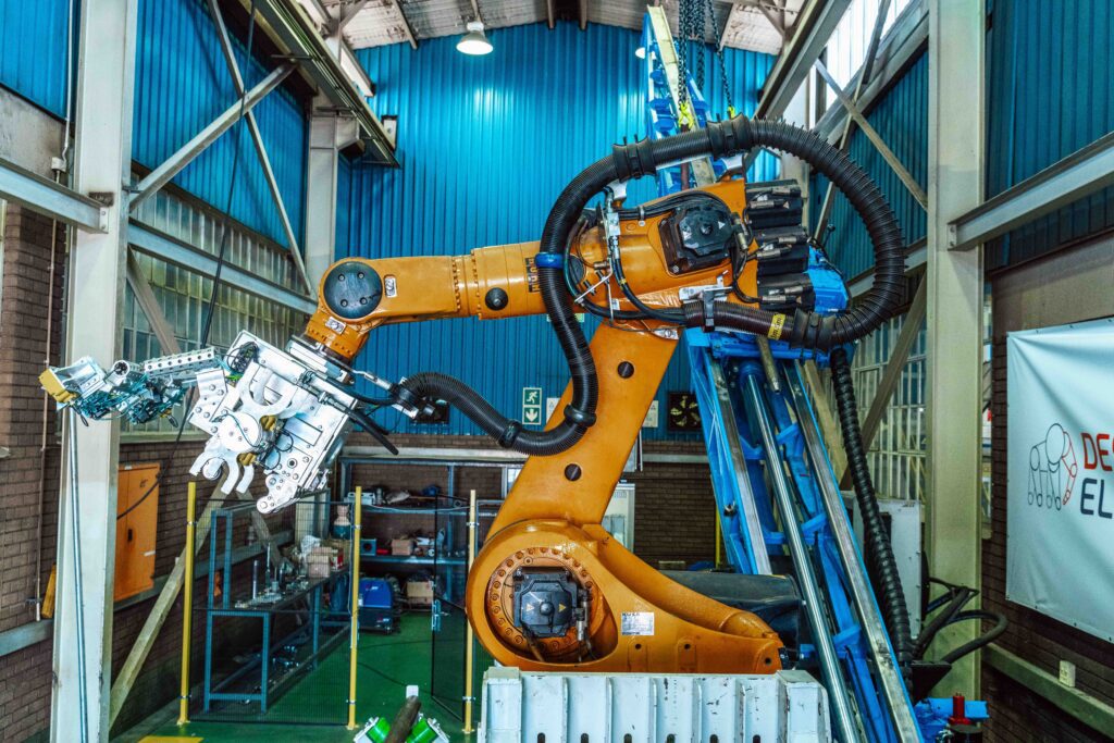 A large industrial robot arm installing mechanical components in a factory setting. The robot is orange and positioned at an angle, surrounded by metal frames and equipment, with a blue wall in the background.